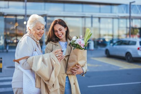 Une dame aux cheveux blancs et une jeune femme rient ensemble en sortant faire des courses, un bouquet de fleurs à la main